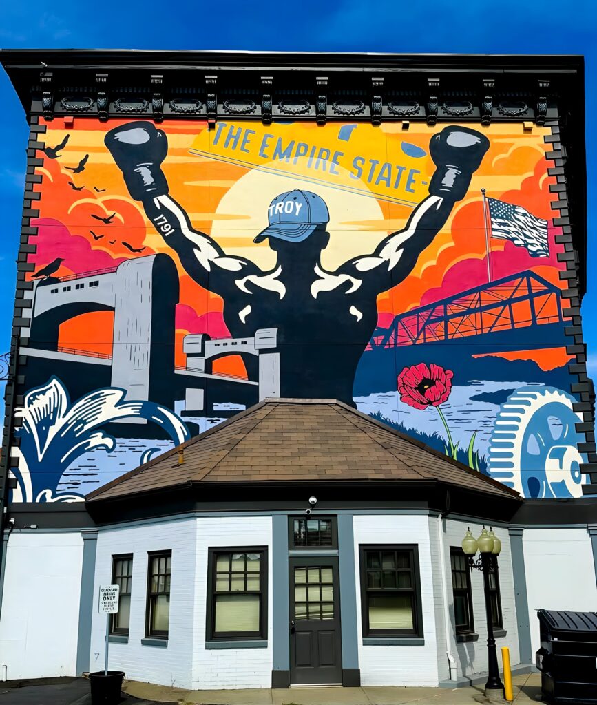 Three-story tall mural on side of building. Central figure is triumphant boxer—arms raised skyward—surrounded by symbols of the city’s heritage, engineering innovation, and patriotic pride.